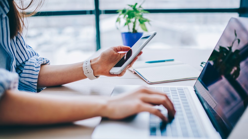 A woman looking at a smartphone while using a laptop