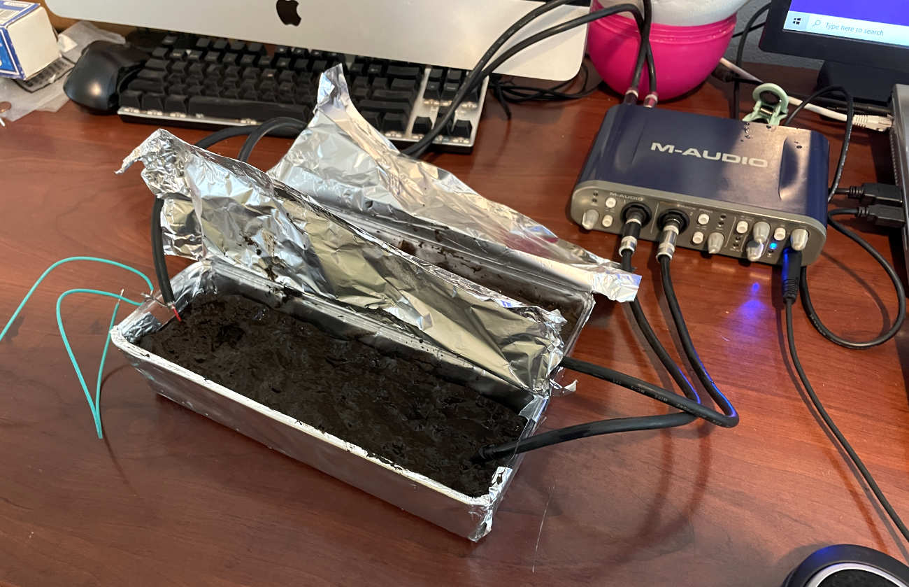 A wide shot of a workspace showing a foil tray of mud connected by DIY cables to a blue M-Audio Fast Track Pro interface, positioned in front of a computer monitor and keyboard.