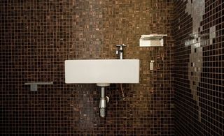 Bathroom with tiles in brown shades. White bathroom sink with silver tap and pipes against the wall.