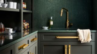 Dark green kitchen with fluted splashback, brass tap and handles, open shelving, and a green stone worktop with a towel draped over the sink.
