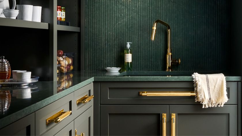 Dark green kitchen with fluted splashback, brass tap and handles, open shelving, and a green stone worktop with a towel draped over the sink.