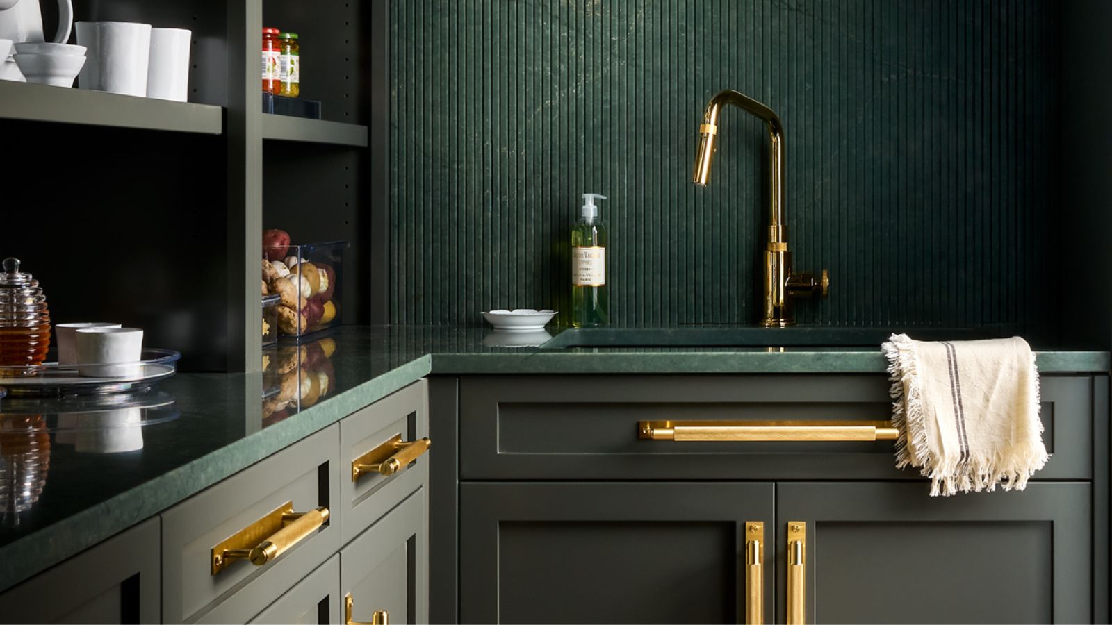Dark green kitchen with fluted splashback, brass tap and handles, open shelving, and a green stone worktop with a towel draped over the sink.