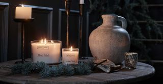 The White Company Nordic Woods signature scent candles on a table in the darkness with candle light and an earthenware jug