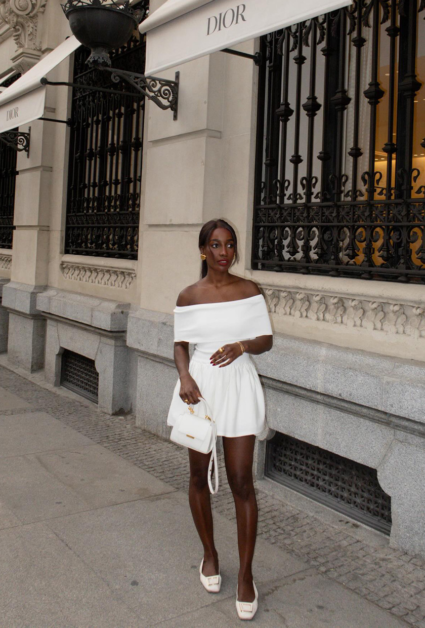 A woman's white dress outfit styled with white slingbacks, a white handbag, and gold earrings.