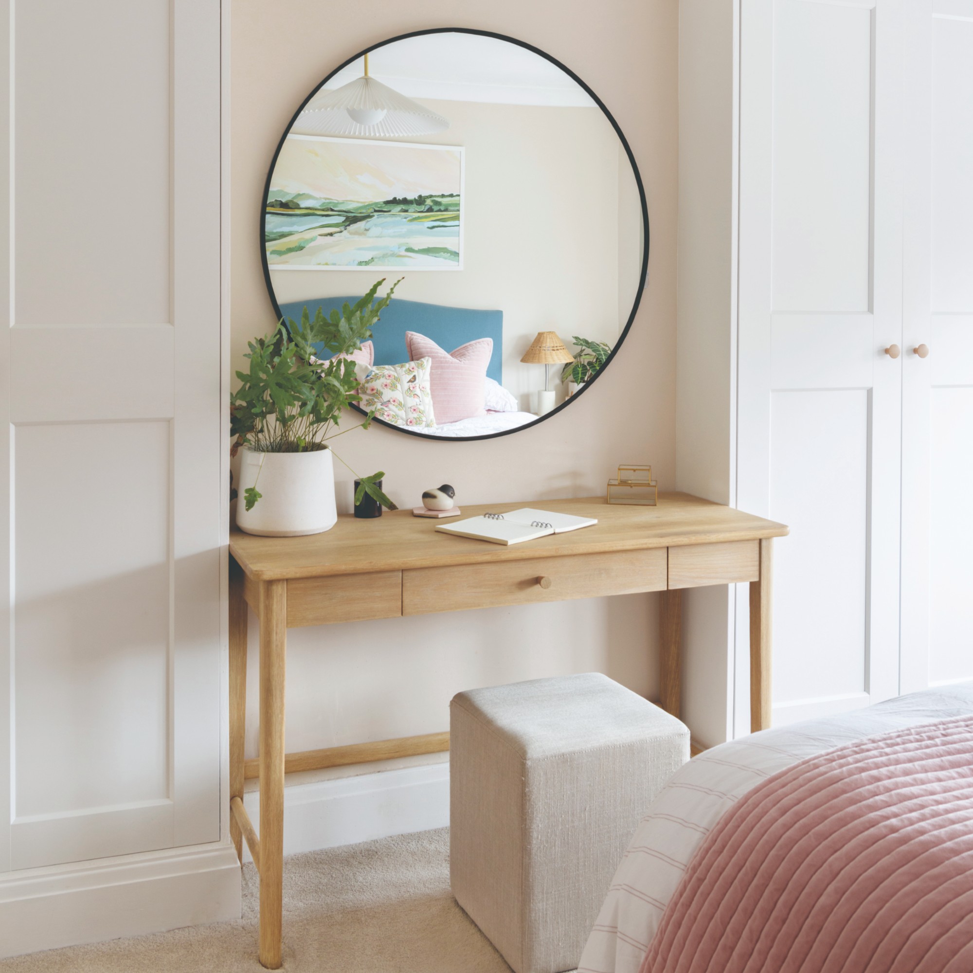 A pastel bedroom with a round mirror hung opposite the bed and reflecting the blue headboard and artwork hung above the bed
