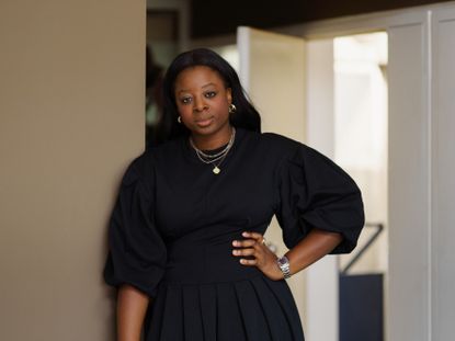 Portraot of Morag Ofili in a black dress standing against a wall in a room 