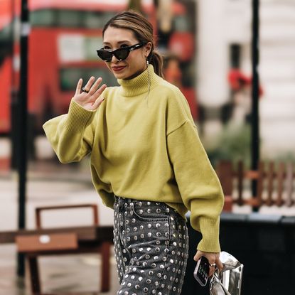 fashion week attendee waving wearing sunglassess, green sweater, and studded jeans
