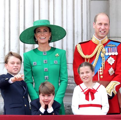 Princess Kate wearing a green coat, Prince William wearing a red military uniform, Prince George wearing a blue suit and pointing, Prince Louis leaning forward with his chin in his hands and Princess Charlotte wearing a white and red dress with a bow