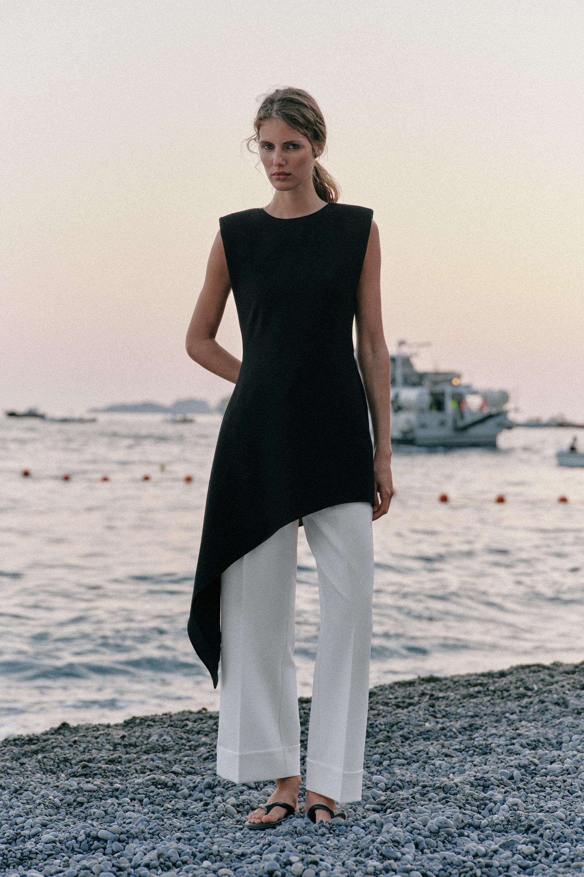 Woman wears black tunic, white pants, and flat sandals.