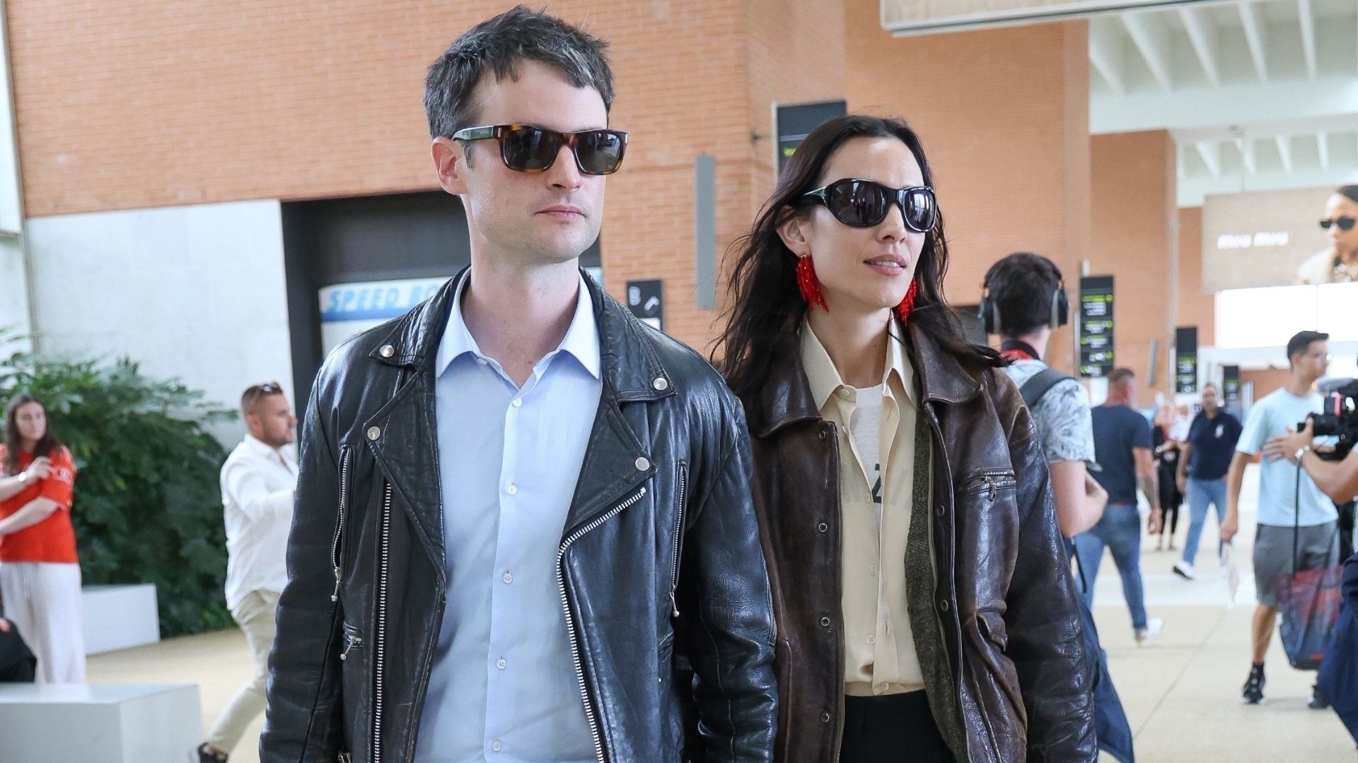 Alexa Chung and Tom Sturidge walk through Venice airport holding hands. Alexa carries a black bag in her hands and wears a brown leather jacket, a yellow shirt, black trousers and black loafers.
