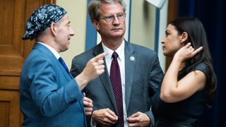 three members of the united states house of representatives talk amongst themselves in a large hearing room