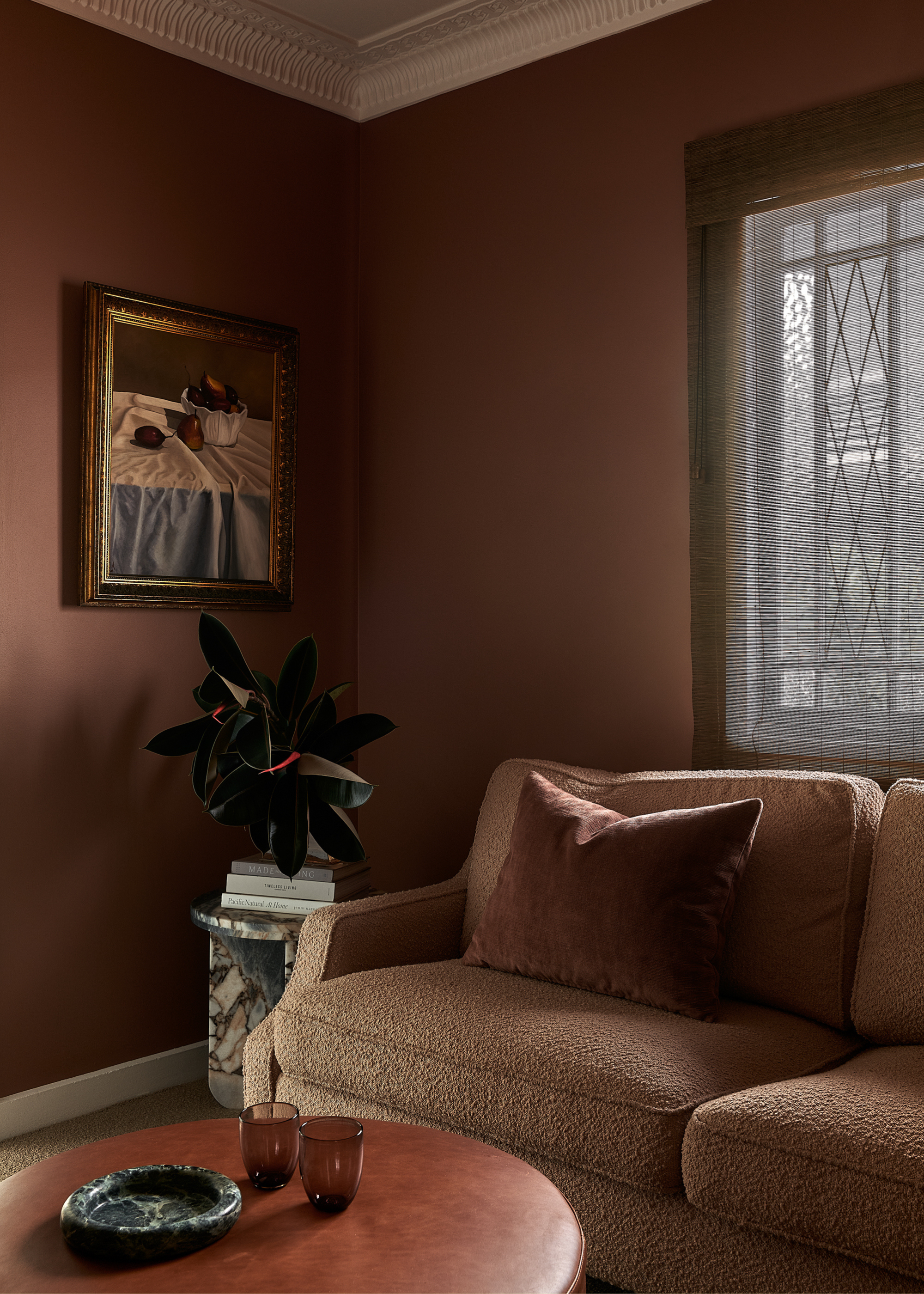 A brown living room corner with a boucle couch, a framed painting, a marble side table with a potted plant, and a round table and a stone tray with a pair of glasses