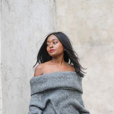 Carrole Sagba wears a gray oversized off shoulder pullover, pale gray suit pants with white feathers on one half leg, black and beige striped leather heels ankle boots, a white matte leather handbag, on April 18, 2021 in Paris, France.