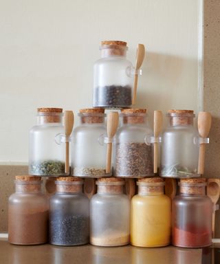 A stack of frosted glass spice jars with colorful spices in them, brown cork lids, and wooden spoons on the handles, on top of a dark brown countertop against a white wall