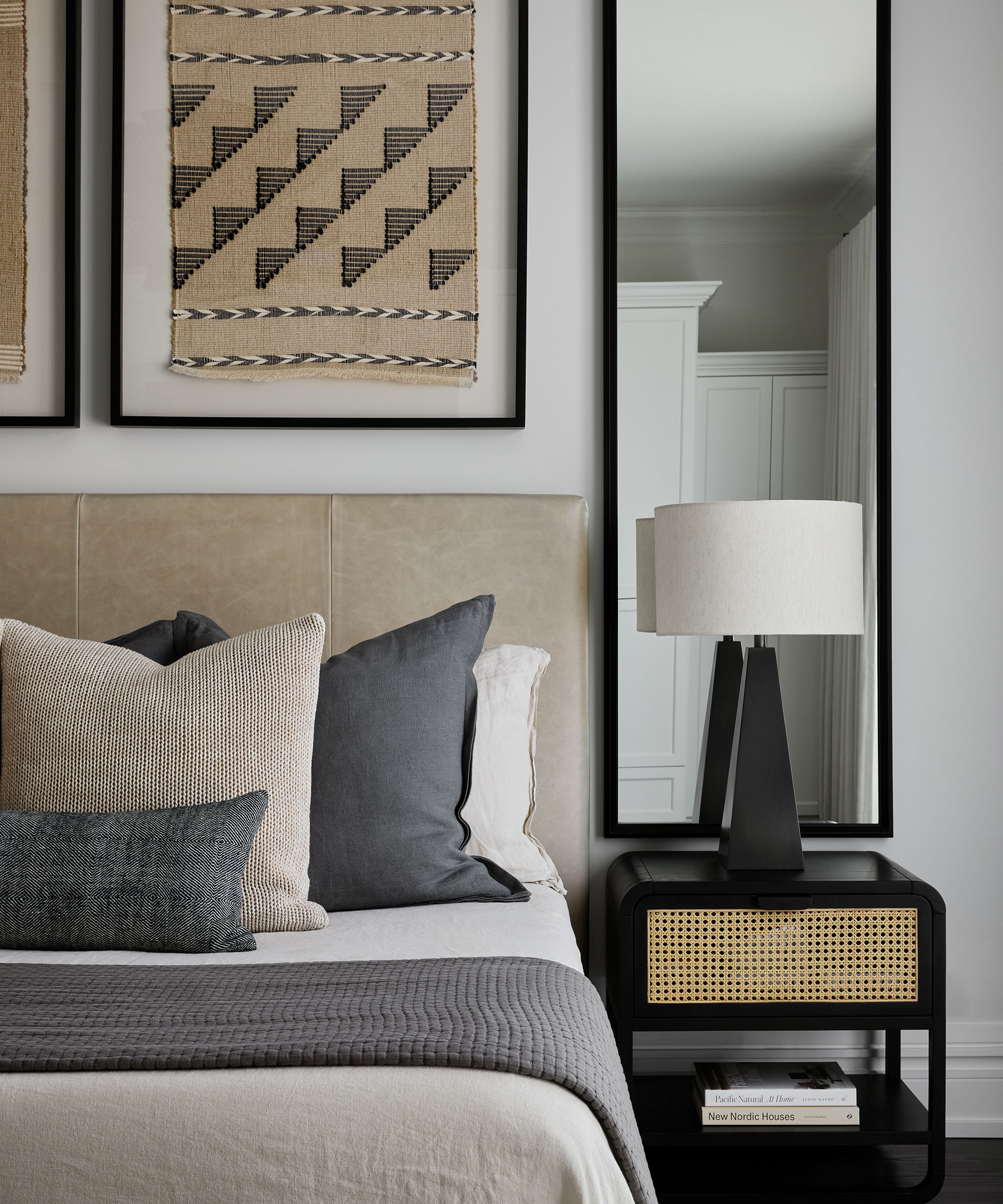 Modern bedroom with artwork and mixture of printed cushions, featuring black nightstand with cane front drawer and curved table lamp with a cream shade