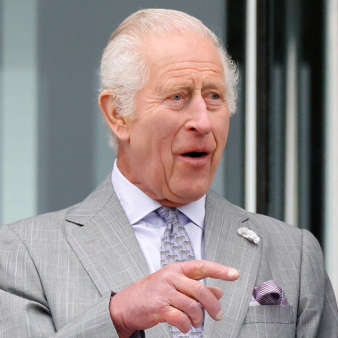 King Charles wears a gray suit and looks shocked while wife Queen Camilla wears a light blue suit jacket