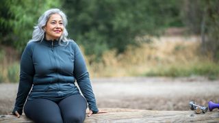 Postmenopausal woman wearing workout clothes sitting on a fallen tree smiling