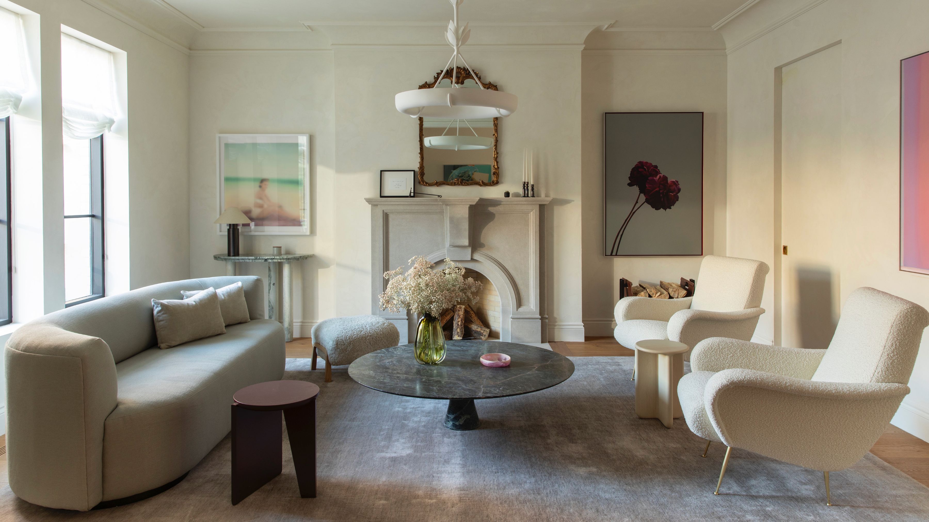 A sophisticated, neutral-toned living room featuring a curved gray sofa, two white boucl&amp;eacute; armchairs, and a large marble coffee table centered around a classic stone fireplace