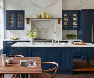 A shaker style kitchen with dark blue cabinets, carefully considered for the storage needs of the owners. The central island has built in cabinets and there are wall mounted cupboards with glass fronts displaying the items within