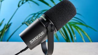 the shure mv7i, a black microphone with a black muff, touch LED pad, XLR connection and TRS input and USB-C input at the rear, photograhped against a blue tom's guide background with a pair of over-ear wired headphones