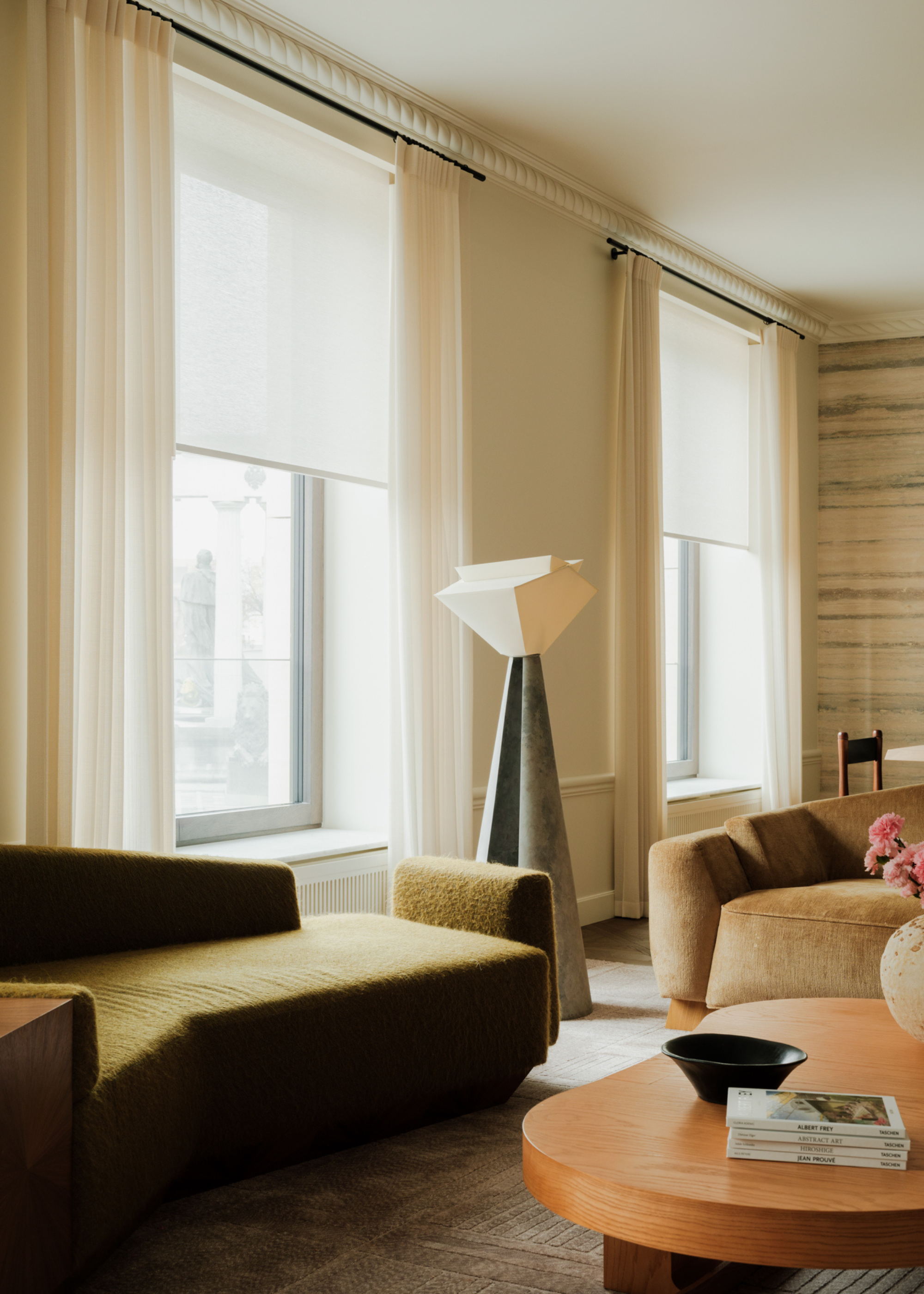 A beautiful living room with a green sofa, a curved wood coffee table, a large floor lamp with a rugged white shade, a vase of flowers, a stack of books, and a bowl