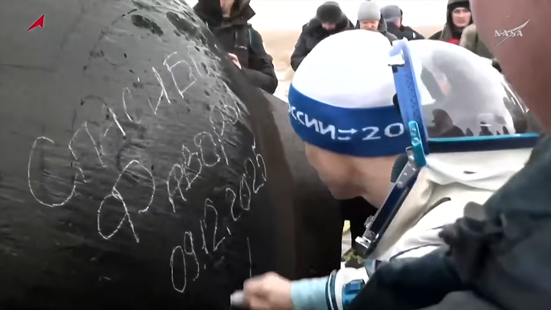 A man in a pressure suit and wool cap uses a piece of chalk to sign his name and the date on the outside of a space capsule