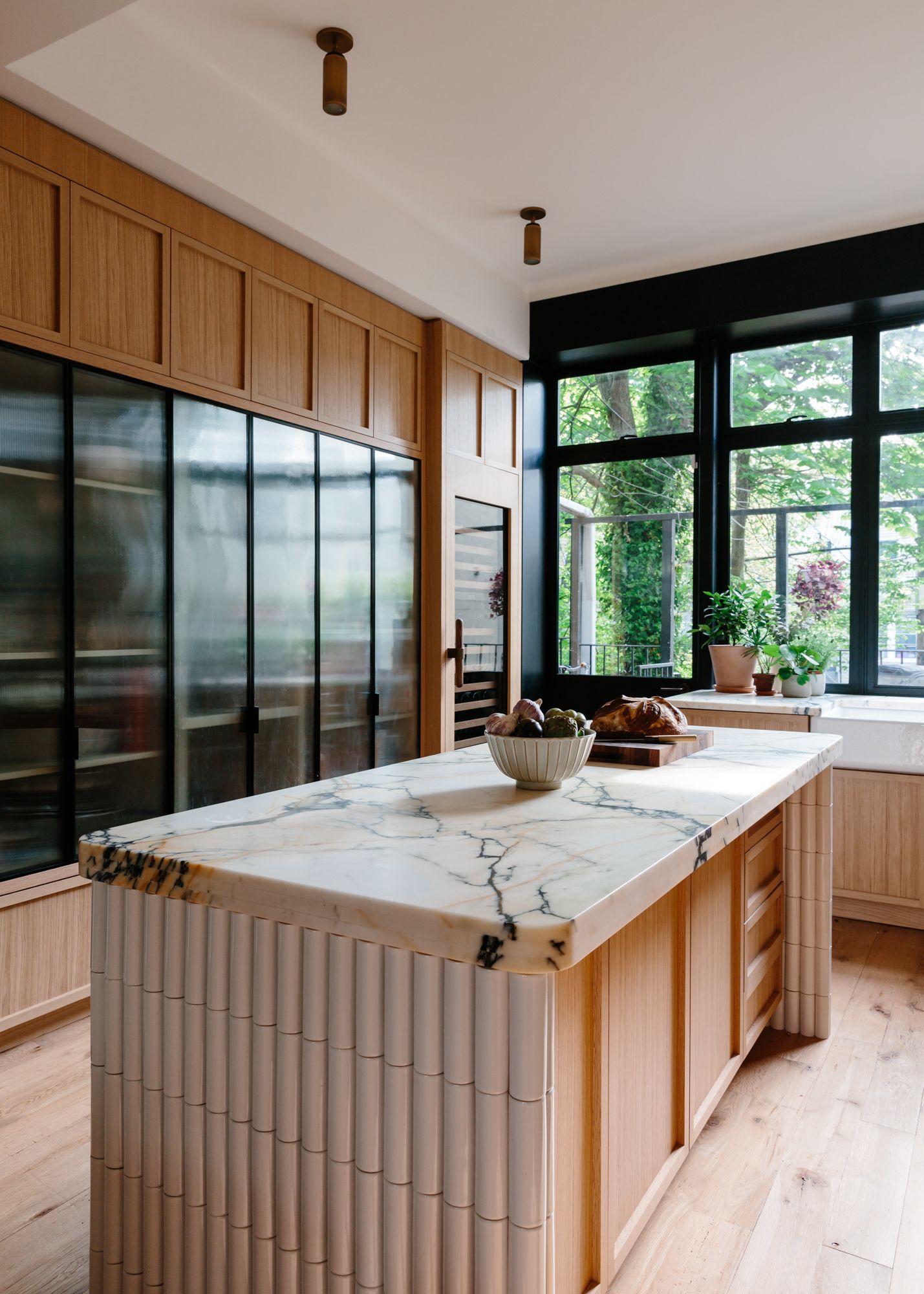A modern kitchen with natural wood joinery, a stone-topped kitchen island and black-framed windows with views of the garden