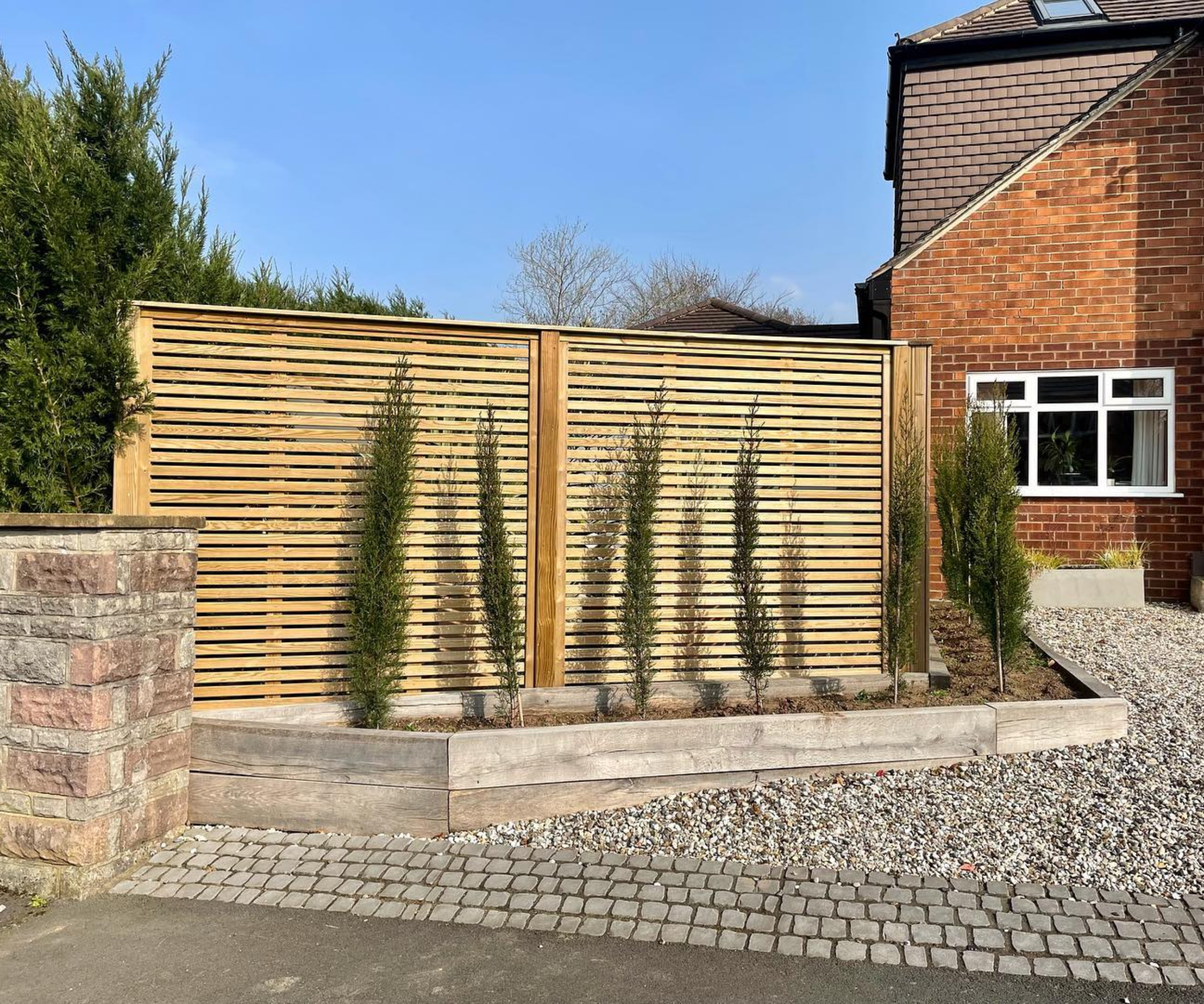 gravel driveway with stone setts before road, driveway fencing idea to one side with small tree planted border in front of fence