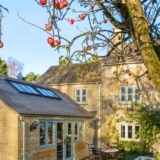 Large beige stone home with sage green windows. There are solar panels on the roof and it is a sunny winters day