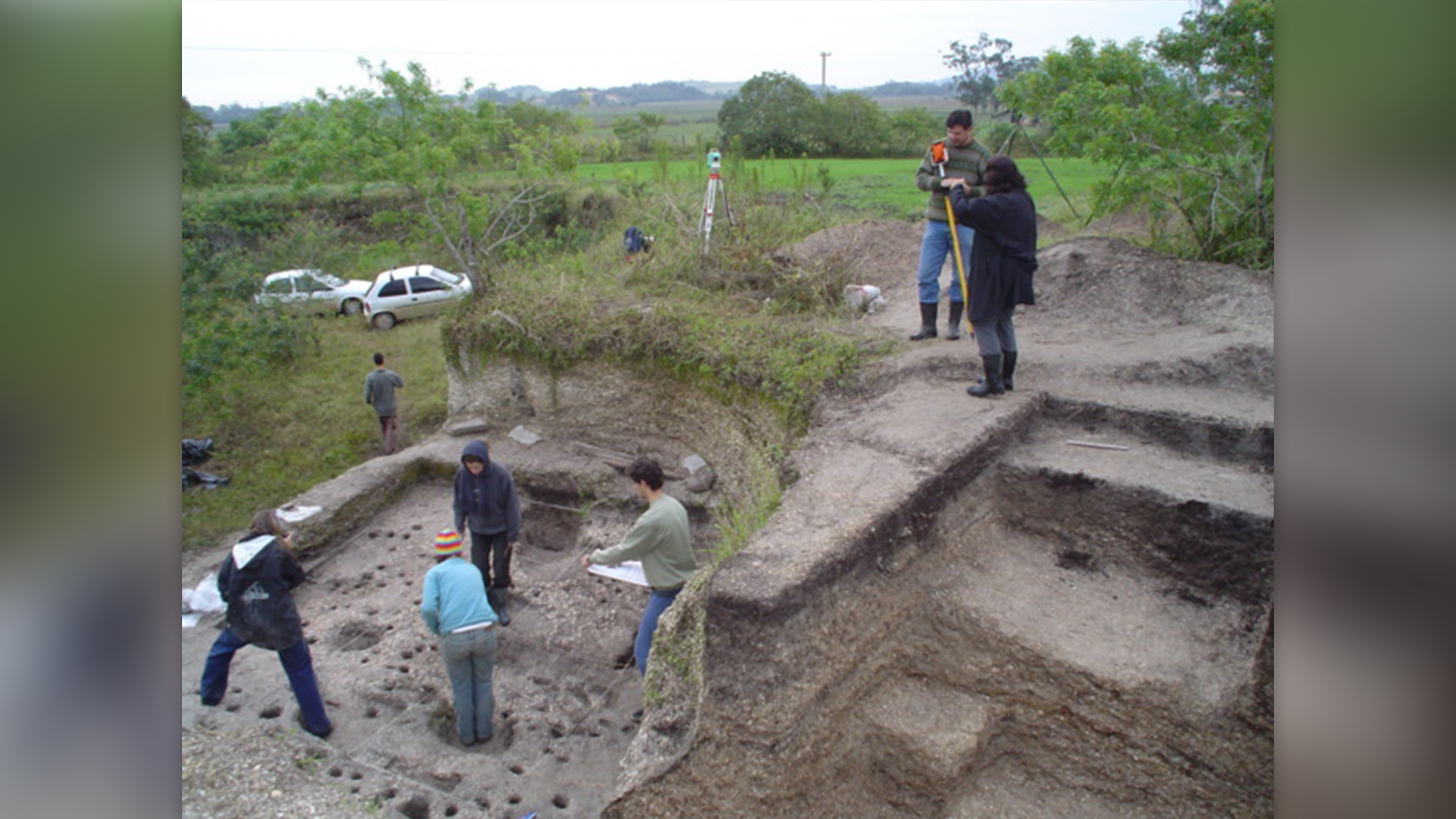 photo shows seven researchers standing around an archeological dig site, excavating structures from the dirt. Greenery and two cars can be seen in the background