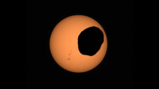 A screenshot from a video showing the Mars moon Phobos crossing the face of the sun. NASA’s Perseverance Mars rover captured the video on April 2, 2022, with its Mastcam-Z camera. You can also spot a group of sunspots on the left.