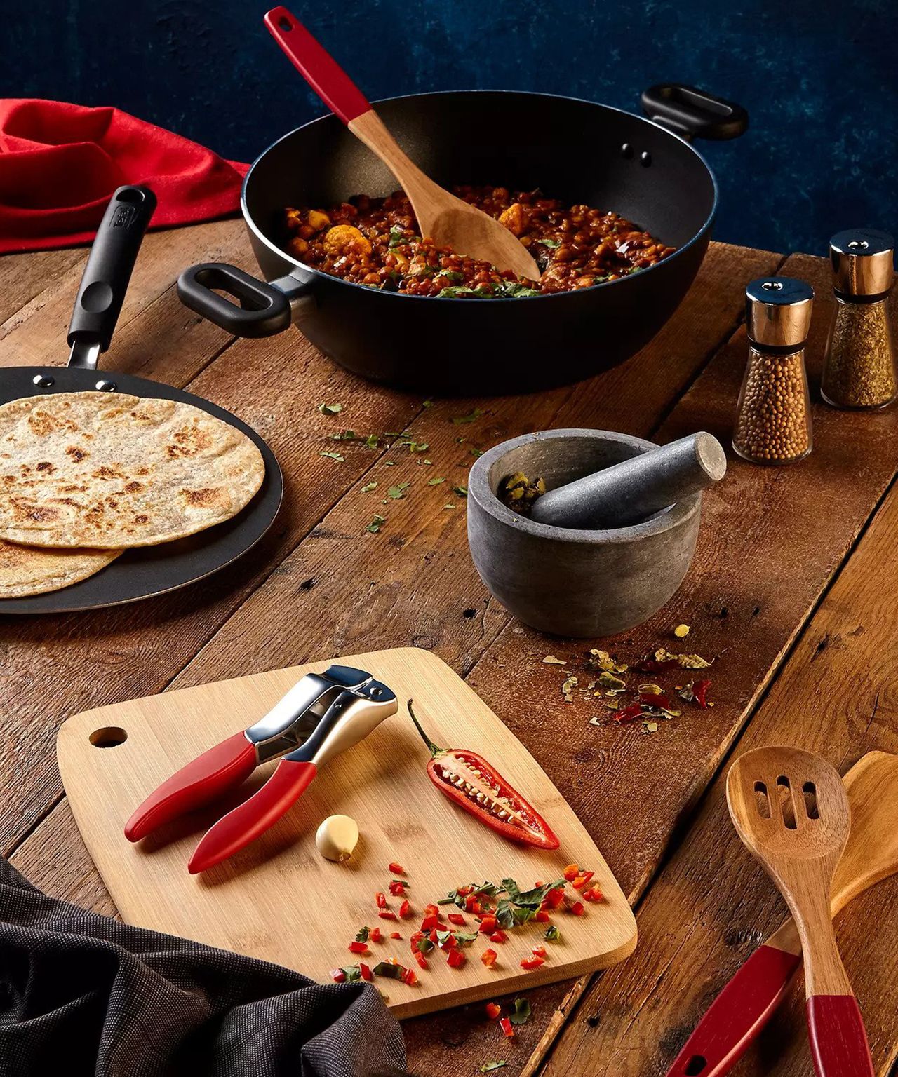 Honed grey marble pestle and mortar on table with cooked dish in pan, cut chilli, garlic with crusher and wooden chopping board