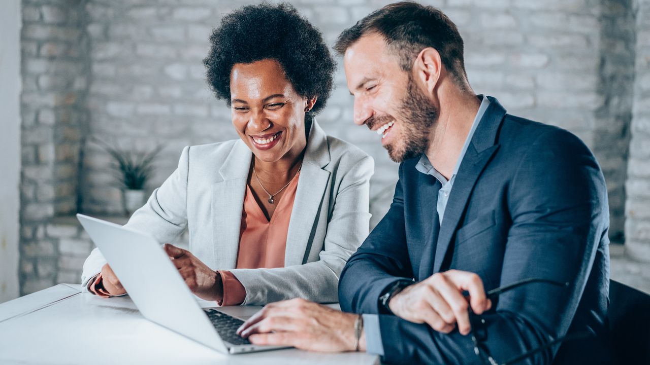 A businesswoman and businessman smile as they look at a laptop together in an office.