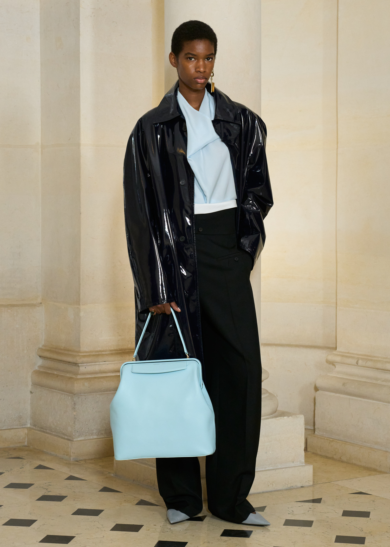 A model in the Carven S/S 26 show wearing a patent-leather coat, a light-blue halter top, trousers, and a light-blue kiss-lock bag.