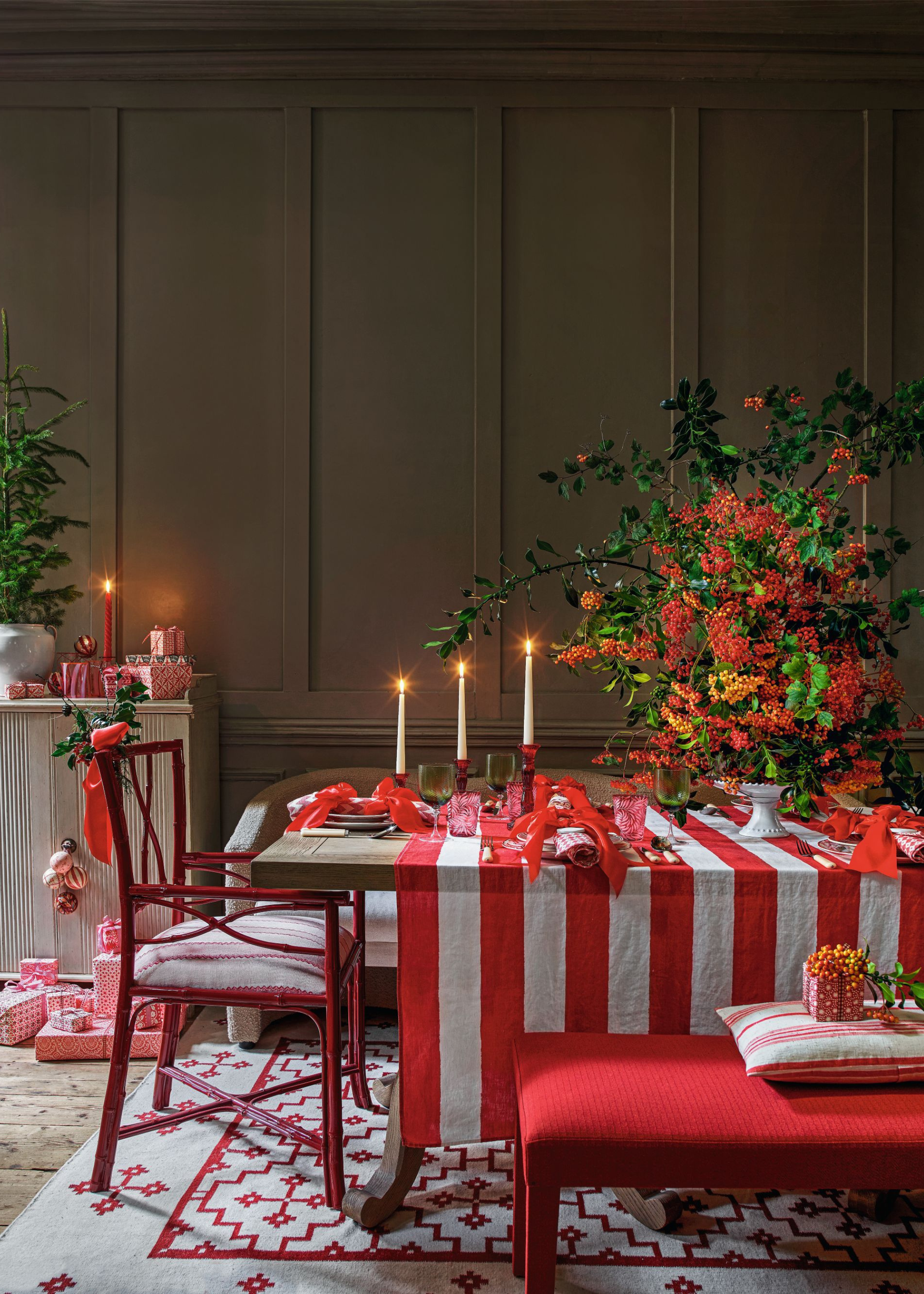 A Christmas table decorated with a red and white striped tablecloth, lit taper candles in varied height red taper candleholders, red napkins and a vase with oversized colorful foliage