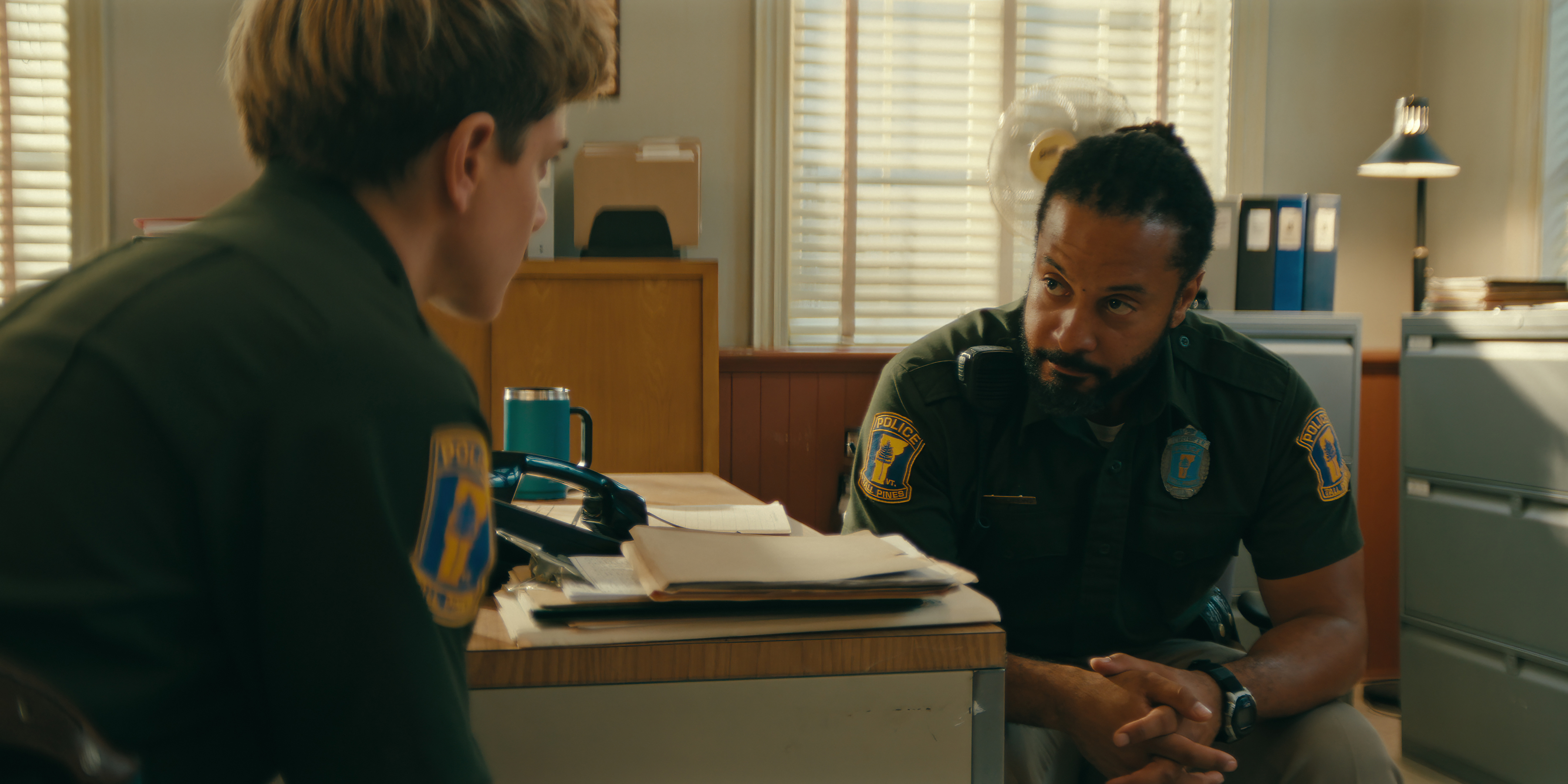 Mae Martin as Alex Dempsey and Brandon Jay McLaren as Dwayne Andrews in cop uniforms in a police office in wayward