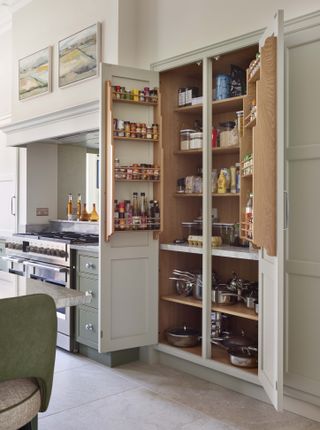 neutral kitchen with open doors showing custom storage, spices storage in doors, saucepans in cupboards underneath, green cabinetry, stone floor, range cooker, artwork