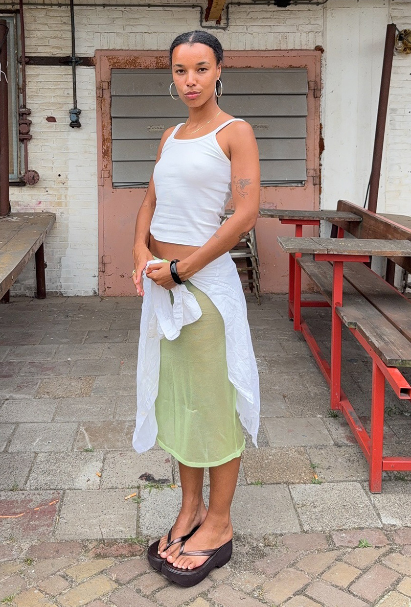The sandal trend wedge flip-flops are shown in a photo of a woman standing outside wearing a white tank top with a sheer green styled styled with a white sarong, silver hoops, and green wedge platform flip-flops