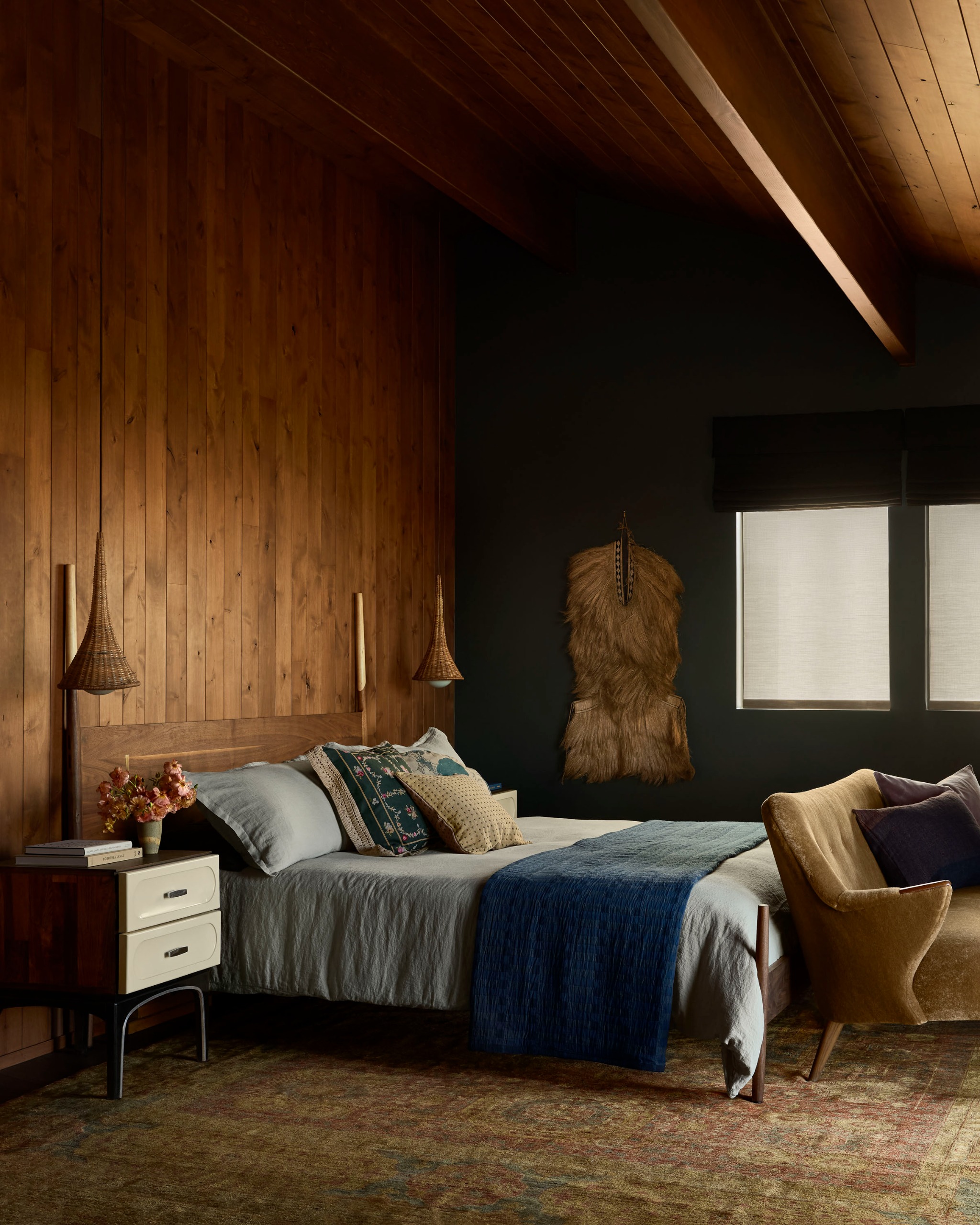 A cozy bedroom with wood paneling, dark green walls, and blue bedding on the bed with teardrop rattan pendants hanging from the ceiling for bedside lights and a small sofa at the foot of the bed