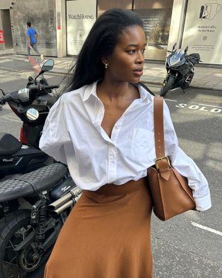 British fashion influencer Marilyn Nwawulor-Kazemaks poses on a sidewalk wearing a white button-down shirt, tan shoulder bag, and camel skirt.