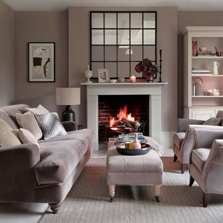neutral living room with pinkish tones, with a burning fireplace and a pink plush sofa, armchair and foot stool set