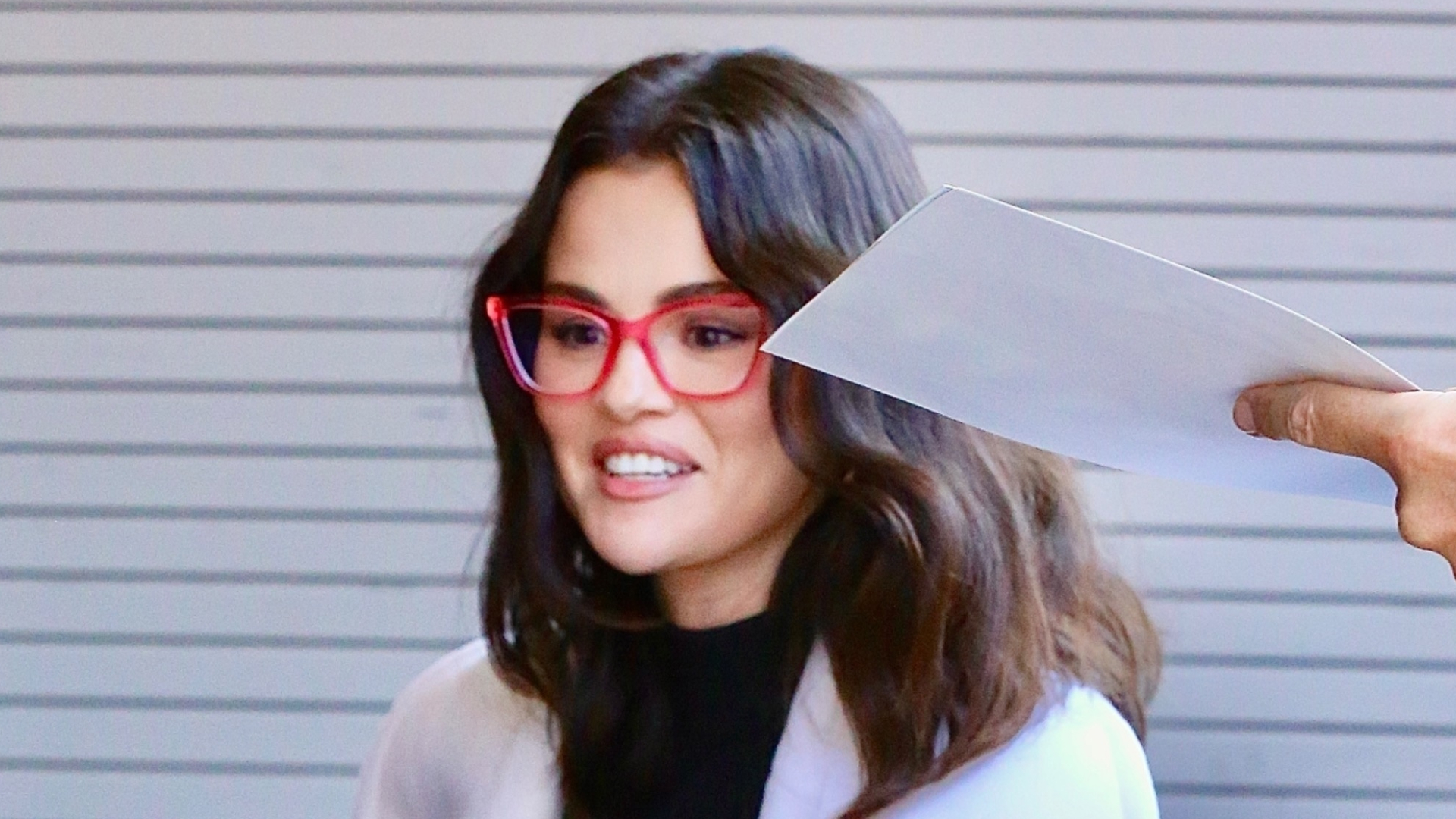 Selena Gomez wearing heeled flip-flops, a black dress, a white coat, and red glasses