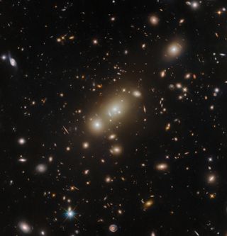 A crowd of oval-shaped elliptical galaxies gather around the largest in the center. They are surrounded by more distant stars and galaxies, that have many shapes and sizes but all are smaller, on a dark background.