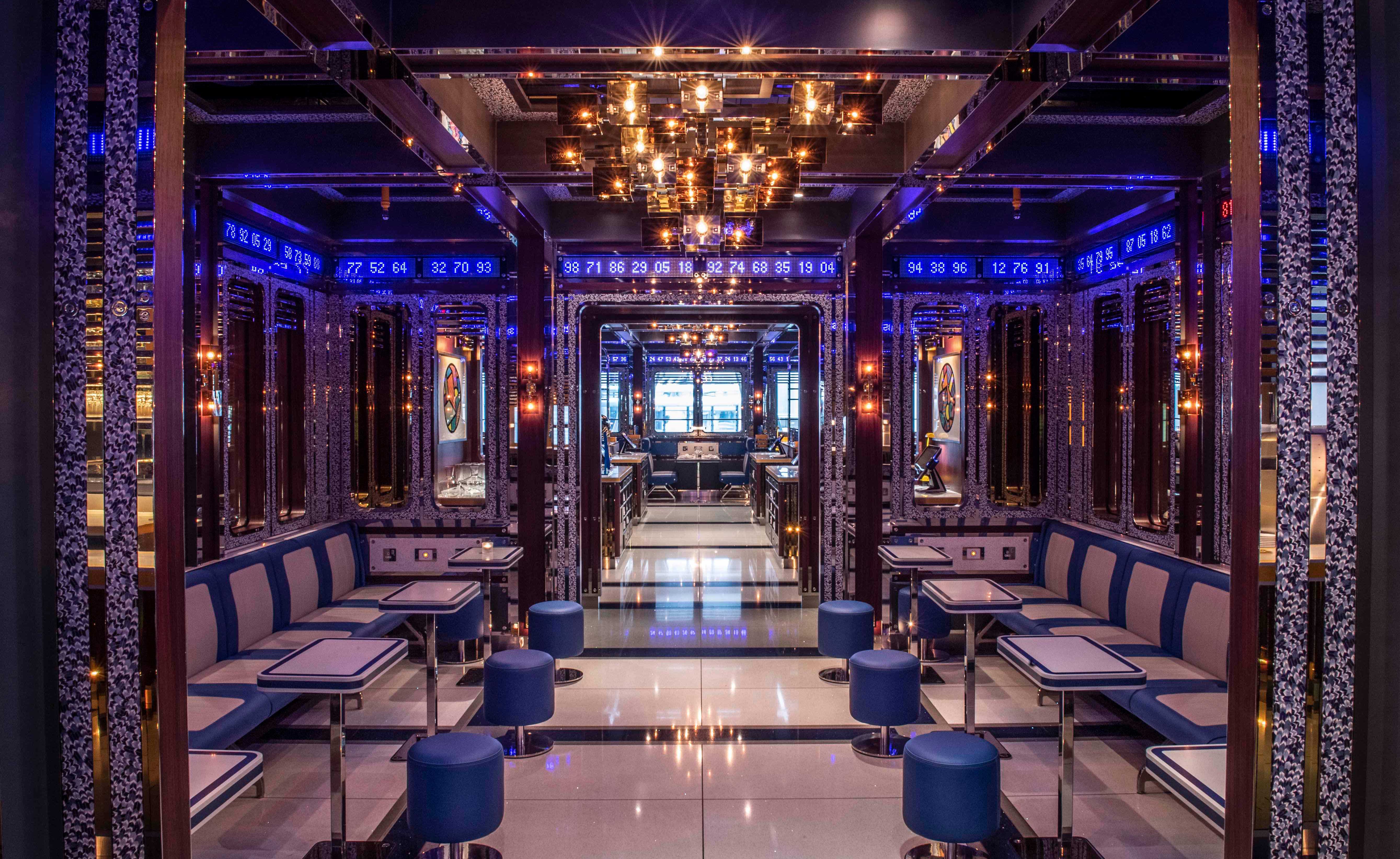 Dining room at Bob Bob Cité restaurant with white and blue tables, white and blue benches against the walls, round blue chairs, wall mirrors, a chandelier and a strip of blue glowing numbers running below the ceiling.