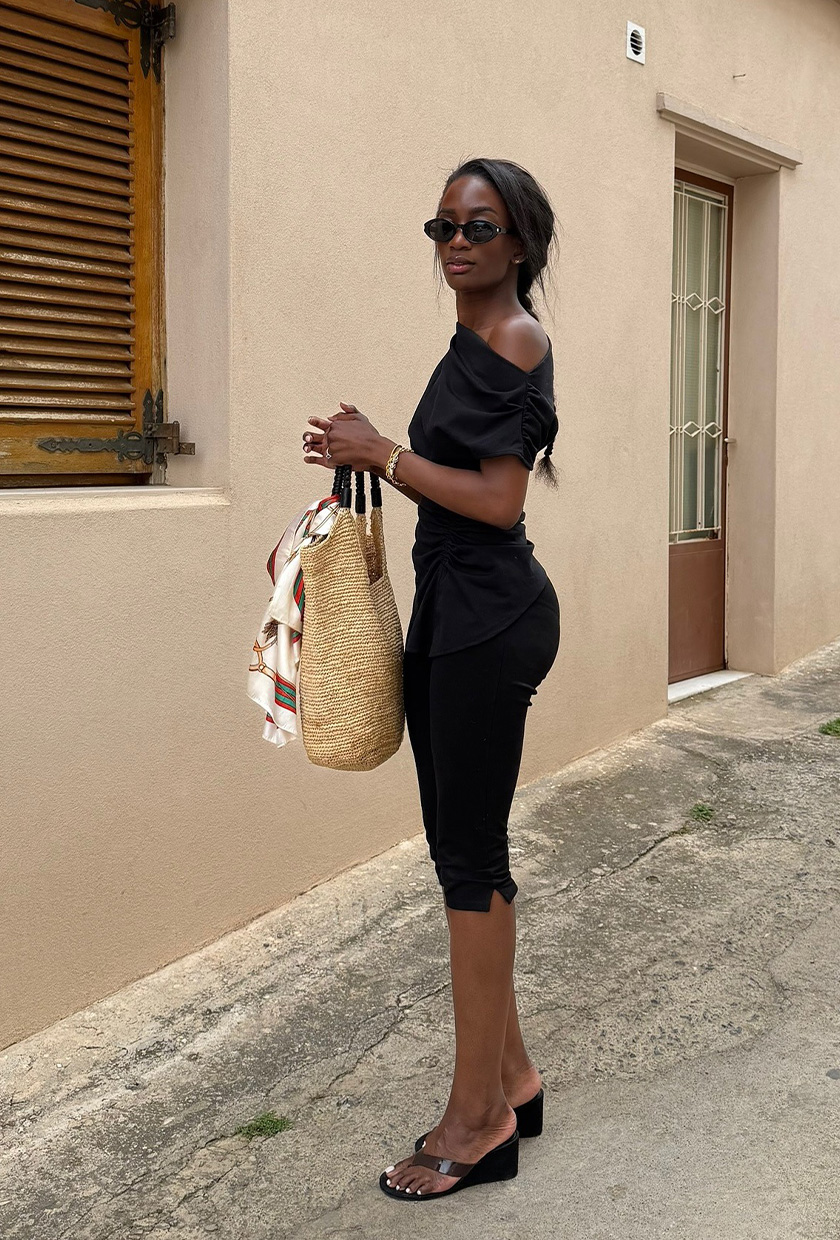 The sandal trend wedge flip-flops are shown in a photo of a woman standing outside wearing a black draped top styled with black petal pushers, black thong wedge shoes, a raffia tote bag, and black sunglasses
