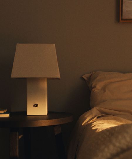IKEA HALGATT cream ceramic lamp sat on a wooden round bedside table, next to a bed with neutral linen bedding.