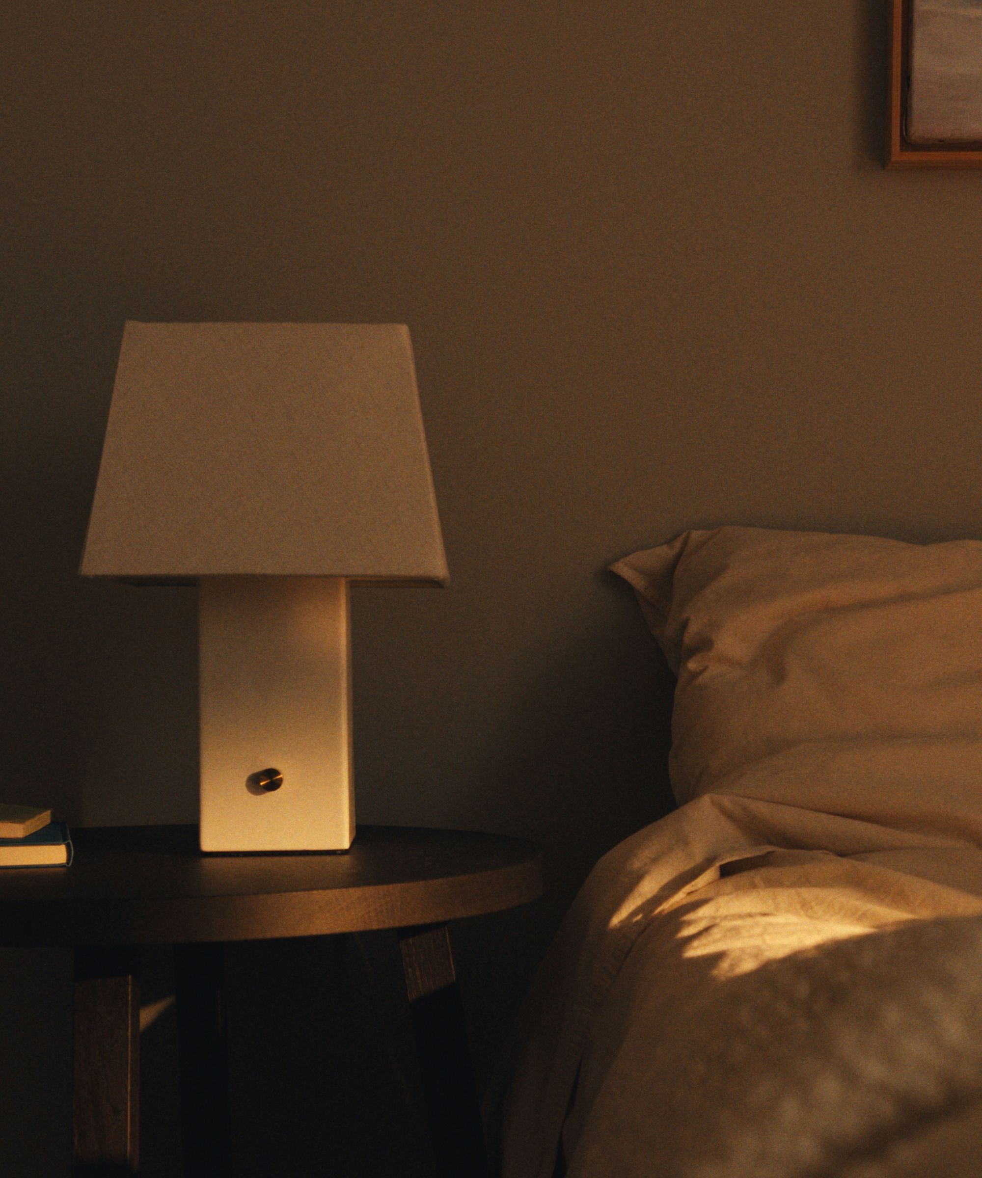 IKEA HALGATT cream ceramic lamp sat on a wooden round bedside table, next to a bed with neutral linen bedding.