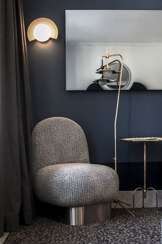 A space age-inspired, chubby and armless armchair in textile and chrome sits atop a mesmerizing blue and grey carpet, next to a gold standing lamp and coffee table, and underneath a spherical gold sconce in a hotel room.