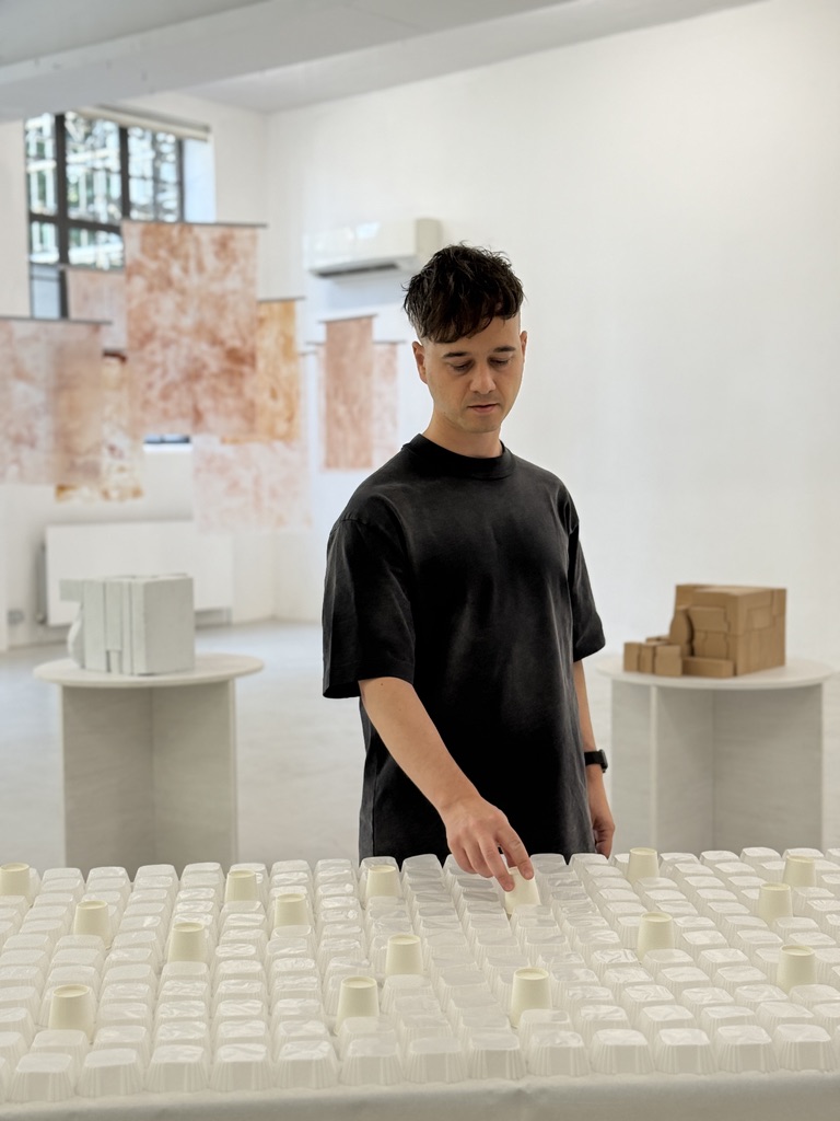 A young male chef dressed in a total black outfit puts the final touches on a food installation showcased in a total-white gallery.