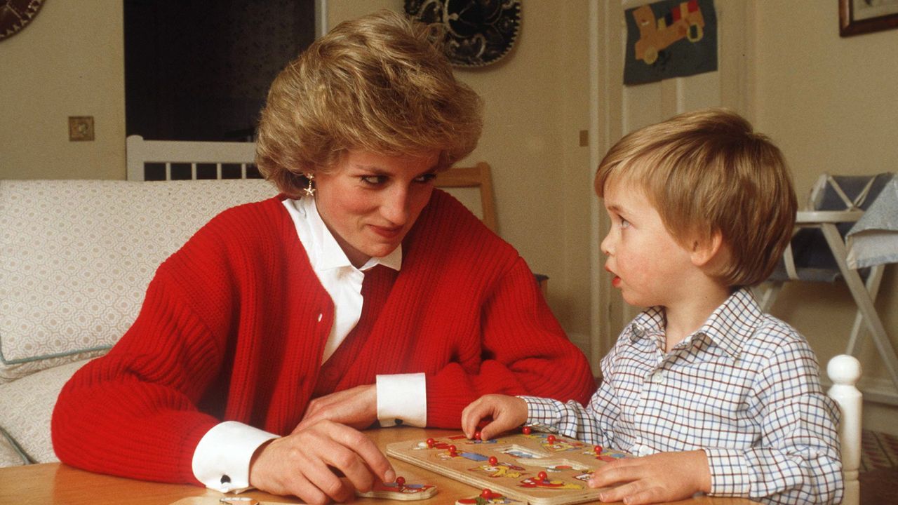 Princess Diana wearing a red cardigan over a white button up shirt sitting at a table playing a puzzle with a toddler Prince William